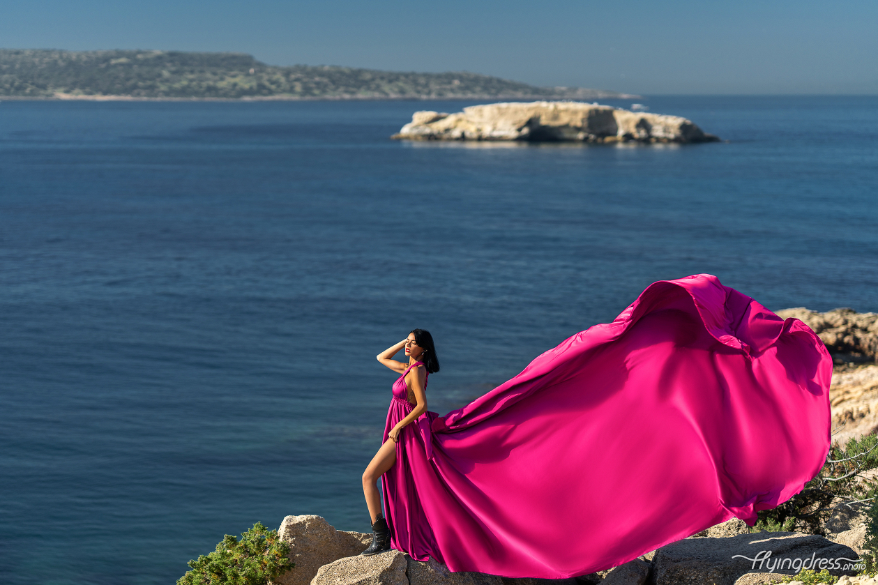 A woman in a flowing fuchsia dress stands confidently on a rocky cliff, with the serene blue waters and distant landforms of Kavouri, Athens in the background.